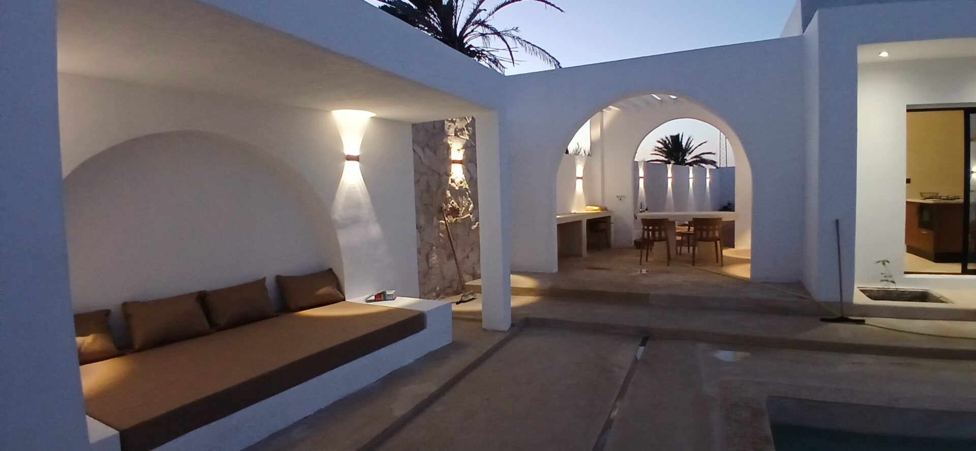 Patio d'une villa Ethic Village Djerba au crépuscule — alcôve voûtée, arches blanches et coin repas sous palmier, ambiance week-end prolongé en couple