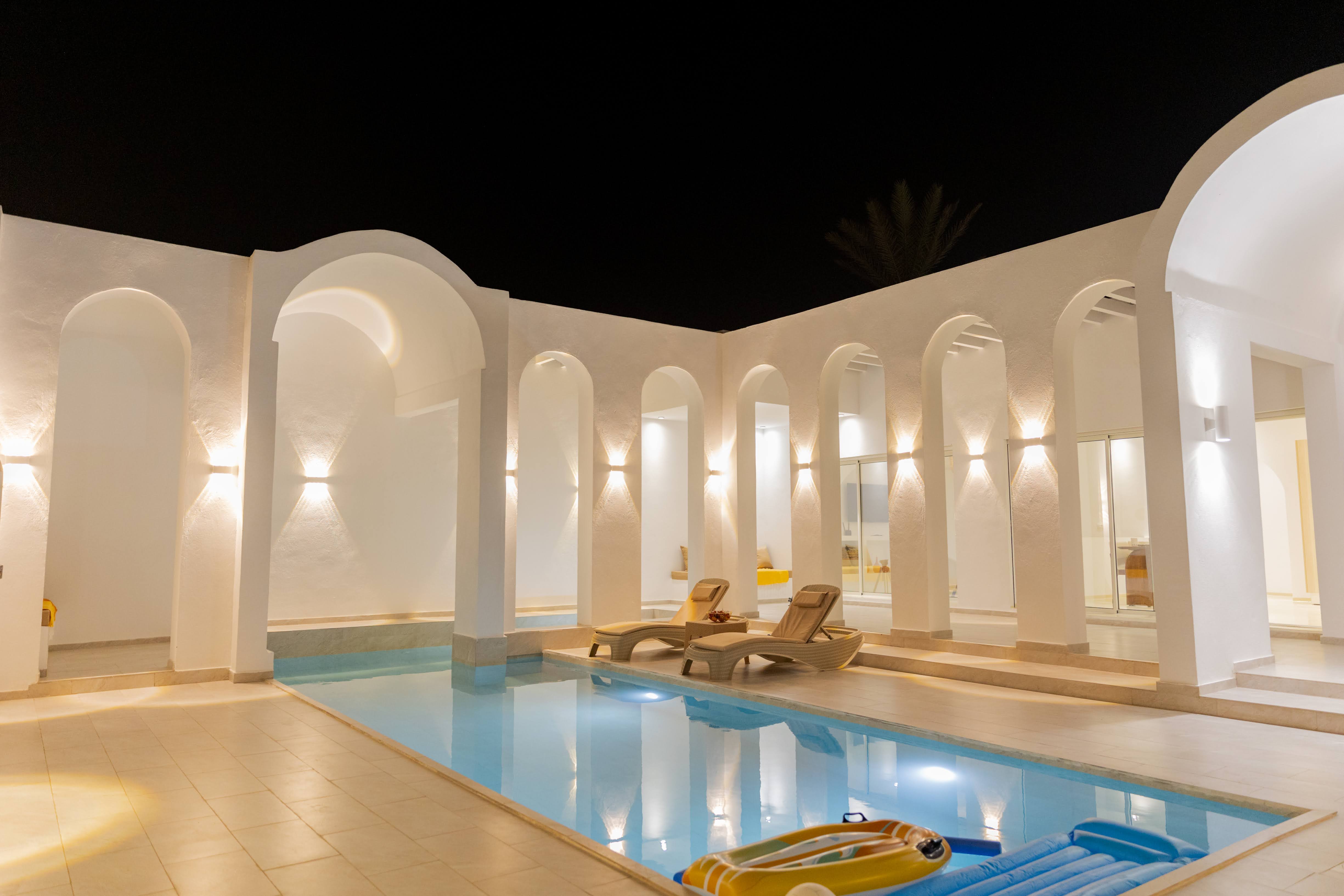Piscine de la villa Jade à Ethic Village Djerba la nuit — arches, transats et éclairage chaud, vue de l'intérieur du jardin clos