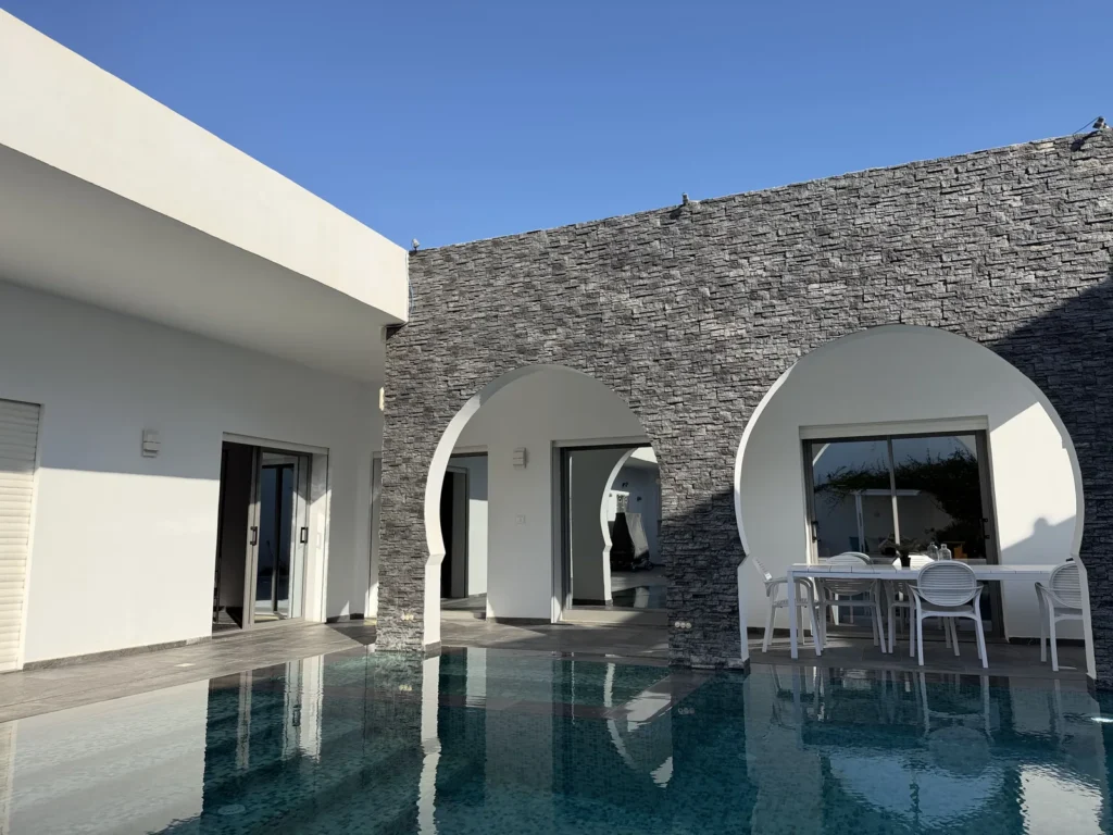 Piscine privée de la villa Diamant à Djerba en octobre, devant un mur de pierre grise percé d'arches blanches, sous le ciel bleu