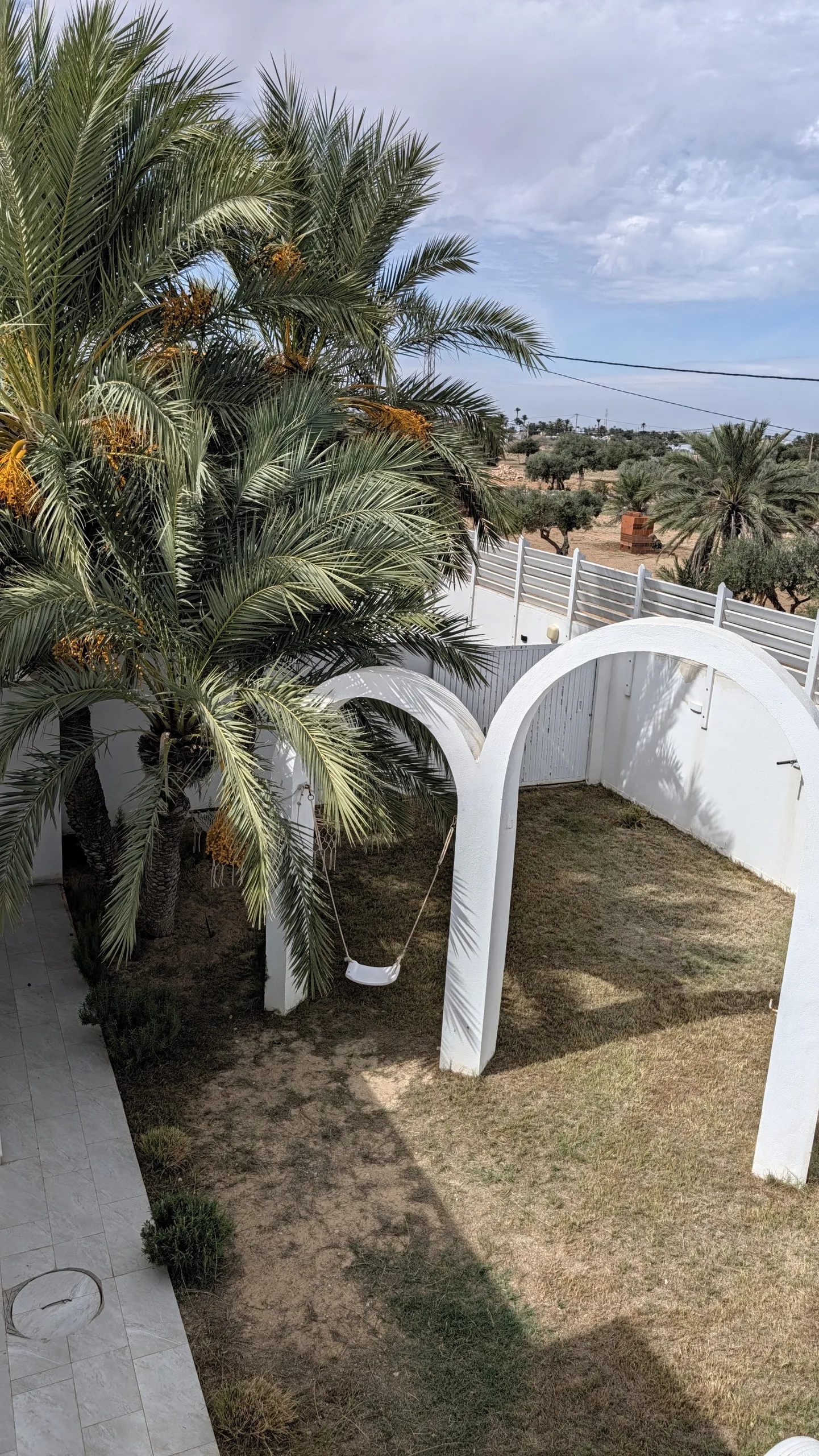 Giardino della villa Jade a Djerba: palme da dattero cariche di frutti, archi bianchi e altalena sotto un cielo sereno