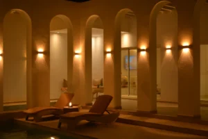 Deux chaises longues et une lanterne allumée au bord de la piscine, sous les arches éclairées de la villa Jade, le soir