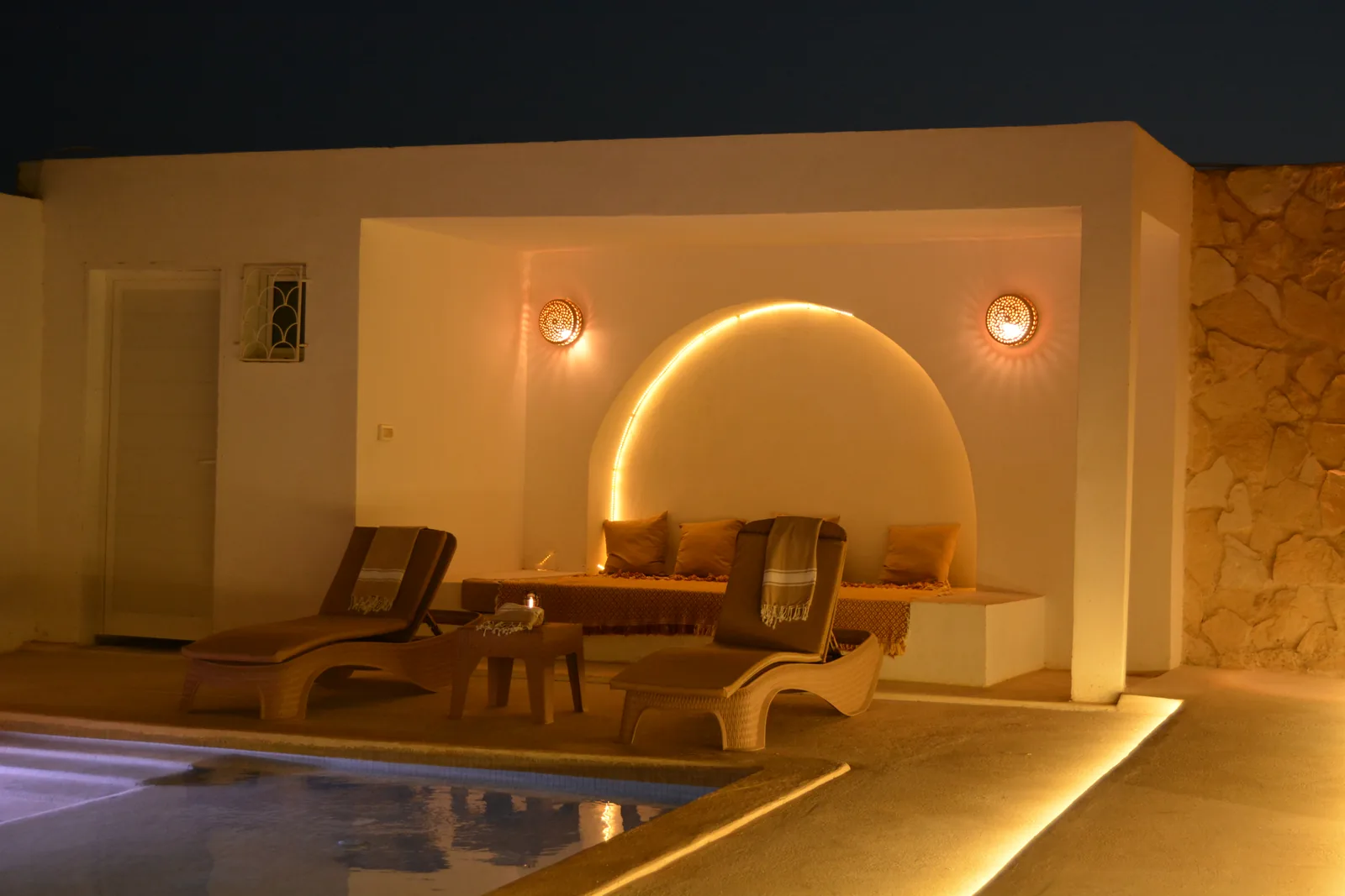 Lit arched alcove and two loungers by the pool of Villa Opale in the evening